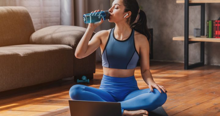 Fit woman in sportswear sitting on mat and drinking from bottle of water while using laptop at home living room